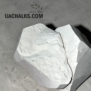 May include: A large, irregular chunk of white chalk with a rough, textured surface. The text UACHALKS.COM and a small leaf icon are visible in the upper portion of the image, indicating the brand name associated with this white mineral product.