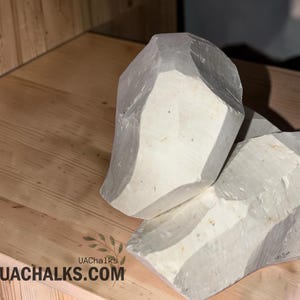 May include: Two large, irregular chunks of off-white chalk with a matte, textured surface. The chalk pieces feature sharp, angular edges and a solid, opaque appearance. The text UAChalks.com is printed in black near the bottom left corner.