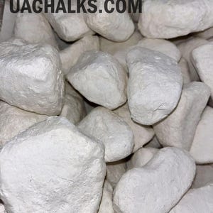 May include: A collection of irregular, matte white chalk chunks with a rough, porous texture. The text UACHALKS.COM is printed in black across the top of the pile, accompanied by a small graphic of a leafy branch above the brand name.