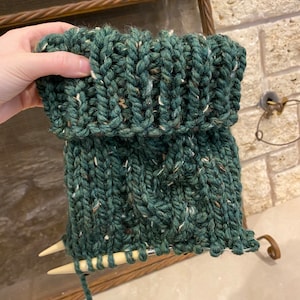 May include: A green knitted cowl with a cable stitch pattern. The cowl is being held by a hand with knitting needles.