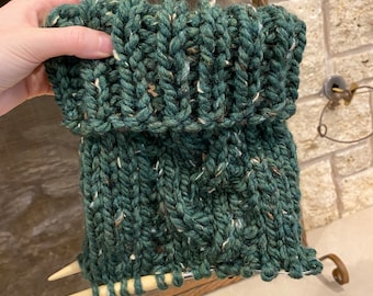 May include: A green knitted cowl with a cable stitch pattern. The cowl is being held by a hand with knitting needles.