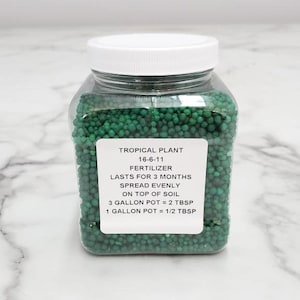 Pode incluir: Um frasco de pl&aacute;stico transparente cheio de gr&acirc;nulos de fertilizante verde. O r&oacute;tulo diz "Tropical Plant 16-6-11 Fertilizante Dura 3 Meses Espalhe uniformemente no topo do solo 3 Gal&otilde;es de vaso = 2 colheres de sopa 1 Gal&atilde;o de vaso = 1/2 colher de sopa"