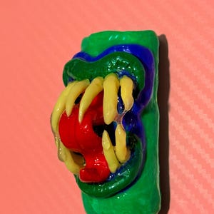 May include: A cylindrical green object featuring a sculpted mouth with prominent yellow fangs and a bright red tongue. The mouth area is outlined in dark blue, and the entire piece has a glossy, smooth finish with a distinct three-dimensional texture.