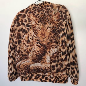 May include: A brown and tan leopard print jacket with a large leopard face design on the front. The jacket is hanging on a black hanger against a white wall. The jacket has long sleeves and a round collar.