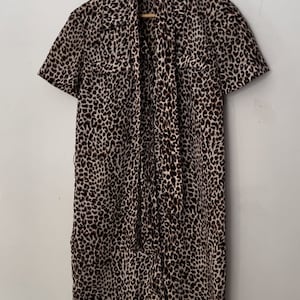 Puede incluir: Un vestido de estampado de leopardo marrón y blanco con manga corta y un lazo en el cuello. El vestido está colgado en una percha de madera.
