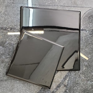 Puede incluir: Dos azulejos cuadrados de cerámica negros, uno ligeramente más pequeño que el otro, con un acabado brillante. Los azulejos están sobre una superficie gris texturizada.