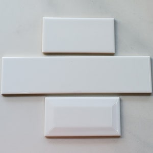 Puede incluir: Tres azulejos de cerámica blanca, uno rectangular, uno largo y estrecho, y uno ligeramente curvo, dispuestos en forma de espiga.