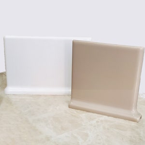 Könnte beinhalten: Zwei keramische Fliesenleisten, eine weiße und eine beige, mit abgerundeter Kante. Die Fliesen liegen auf einer Marmorfläche.