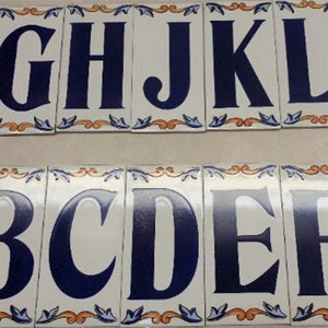 May include: Eight white ceramic tiles with blue letters spelling out the alphabet from B to L. Each tile has a decorative orange and blue border.