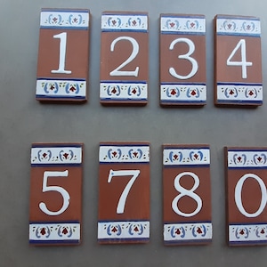 Könnte beinhalten: Satz von zehn Terrakotta-Hausnummern mit weiß lackierten Zahlen und blauen und roten Blumendesigns. Die Zahlen sind 1, 2, 3, 4, 5, 7, 8 und 0.