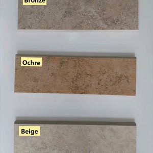 Könnte beinhalten: Drei Keramikfliesenmuster in verschiedenen Brauntönen. Die obere Fliese ist mit "Bronze" beschriftet, die mittlere Fliese mit "Ocker" und die untere Fliese mit "Beige".