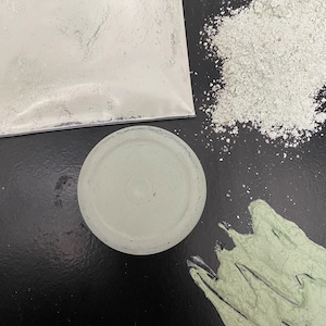 May include: Three containers of white powder, one in a plastic bag, one in a small bowl, and one in a smear on a black surface. The powder is a light green color.