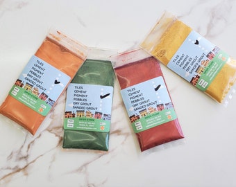 Puede incluir: Cuatro pequeñas bolsas de polvo de pigmento de color para usar en la lechada de baldosas. Las bolsas están etiquetadas "Tiles Cement Pigment Pebbles Dry Grout Sanded Grout" y tienen una pequeña ilustración de casas. Los colores son naranja, verde, rojo y amarillo.