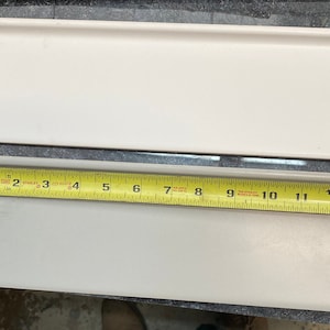 May include: Two white ceramic tiles with rounded edges. The top tile is 12 inches long and the bottom tile is 13 inches long. A yellow measuring tape is used to measure the tiles.