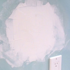 May include: A close-up of a light blue wall with a white patch of drywall compound. A white electrical outlet is visible in the lower right corner of the image.