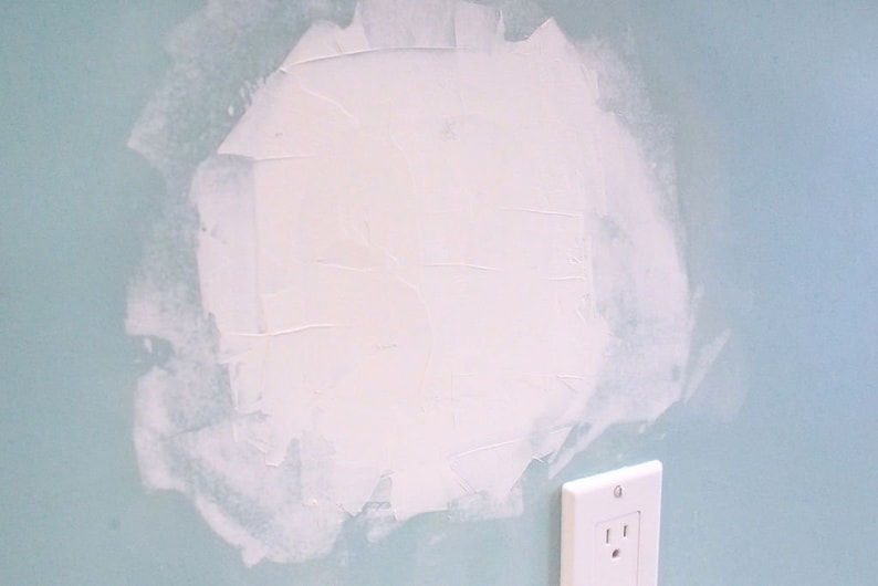 May include: A close-up of a light blue wall with a white patch of drywall compound. A white electrical outlet is visible in the lower right corner of the image.