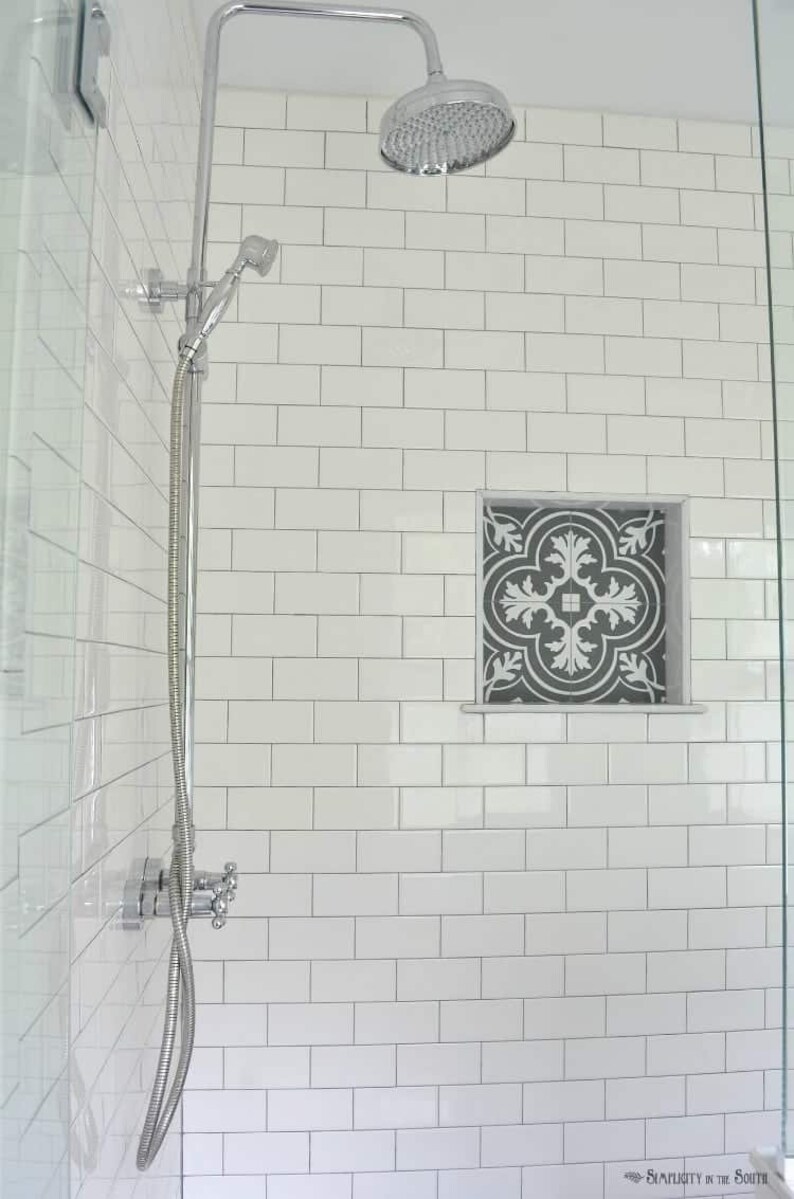 May include: A white tiled shower with a chrome shower head and a decorative tile inset. The shower has a chrome shower arm and a handheld shower head.