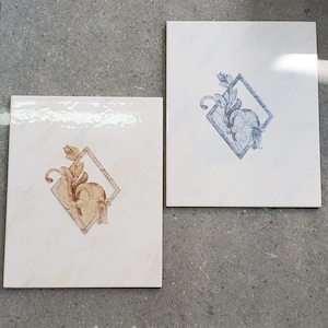 May include: Two white ceramic tiles with a diamond-shaped design. The tile on the left has a gold floral design, while the tile on the right has a blue floral design.