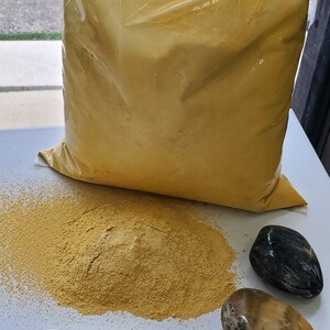 May include: A large bag of yellow grout powder with a smaller pile of grout powder on a white surface. There are two smooth stones, one black and one light brown, next to the pile of grout. A green label with the text "dife pi grout" is on the surface.