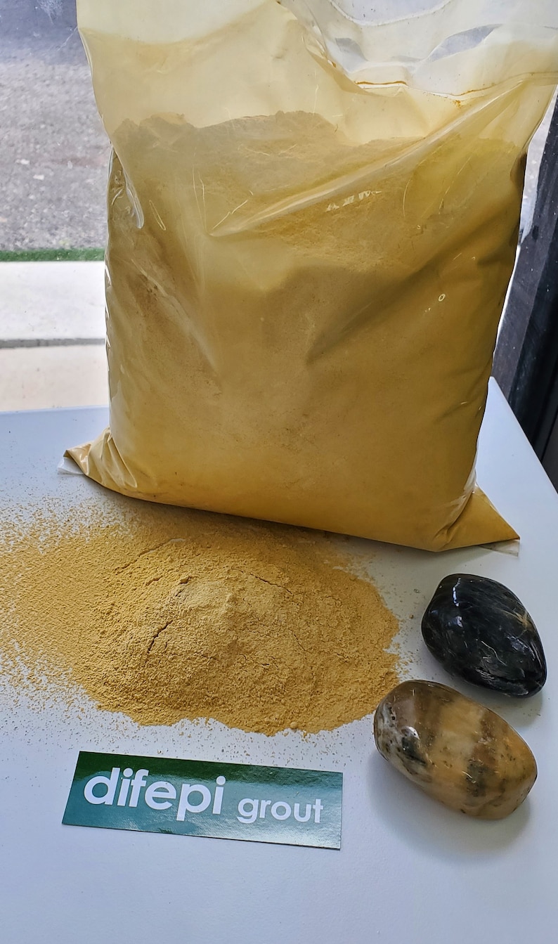 May include: A large bag of yellow grout powder with a smaller pile of grout powder on a white surface. There are two smooth stones, one black and one light brown, next to the pile of grout. A green label with the text "dife pi grout" is on the surface.
