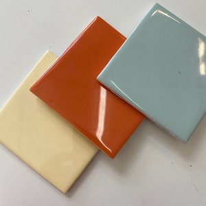 Könnte beinhalten: Drei Keramikfliesen in verschiedenen Farben: ein helles Gelb, ein leuchtendes Orange und ein helles Blau.