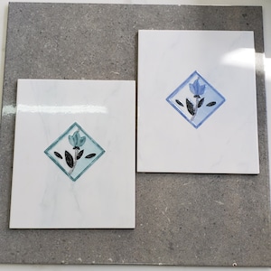 May include: Two white square tiles with a floral design. Each tile features a blue flower within a diamond-shaped frame. The flower has black and grey details. The tiles are on a grey surface.