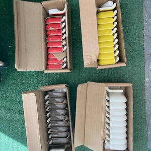 Puede incluir: Cuatro cajas de piezas de moldura de azulejos de cerámica. Las cajas están abiertas y muestran las piezas de moldura de azulejos de cerámica en el interior. Las piezas de moldura de azulejos de cerámica son de cuatro colores diferentes: rojo, amarillo, marrón y blanco.