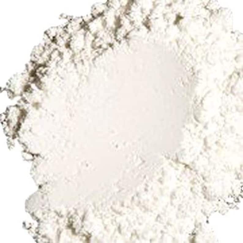 May include: A close-up of a white powder, likely a cosmetic product like face powder or setting powder. The powder is in a pile, with a slightly rough texture.