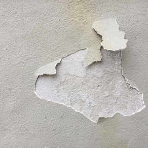 Könnte beinhalten: Nahaufnahme einer grauen, strukturierten Wand mit einem großen Abschnitt abblätternder Farbe. Die abblätternde Farbe zeigt den darunter liegenden Putz.