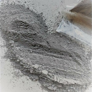 Könnte beinhalten: Nahaufnahme eines Haufens grauen Pulvers auf einer weißen Oberfläche. Das Pulver rieselt aus einer durchsichtigen Plastiktüte.