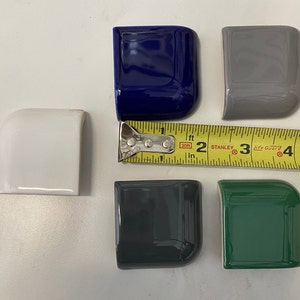Op de afbeelding: Zes keramische tegelhoekstukken in verschillende kleuren, waaronder wit, blauw, grijs, donkergrijs en groen. De tegels zijn gerangschikt op een wit oppervlak met een meetlint.