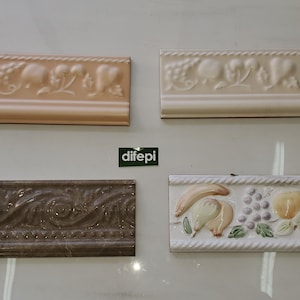 Puede incluir: Cuatro piezas de moldura de azulejos de cerámica con diseños decorativos. Dos son blancas con un patrón floral, una es marrón con un patrón de remolino y una es blanca con un diseño de frutas y uvas.
