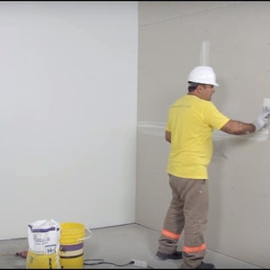 May include: A worker wearing a white hard hat and yellow shirt applies white adhesive to a drywall panel with a trowel. Two buckets of white adhesive are in the foreground.