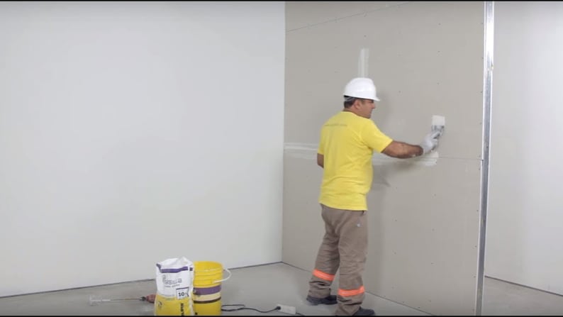 May include: A worker wearing a white hard hat and yellow shirt applies white adhesive to a drywall panel with a trowel. Two buckets of white adhesive are in the foreground.