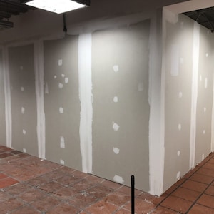 May include: A close-up of a wall with drywall patches and tape. The wall is painted a light gray color. The floor is made of red tiles.
