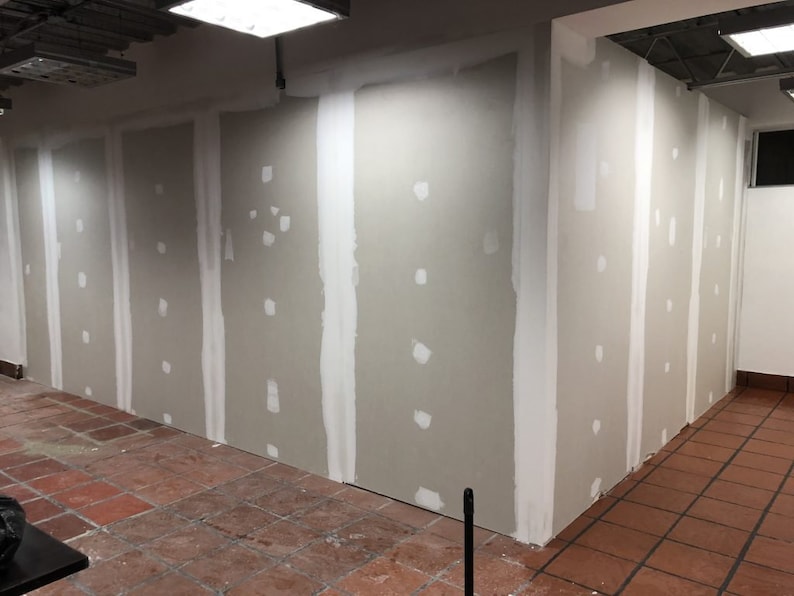 May include: A close-up of a wall with drywall patches and tape. The wall is painted a light gray color. The floor is made of red tiles.