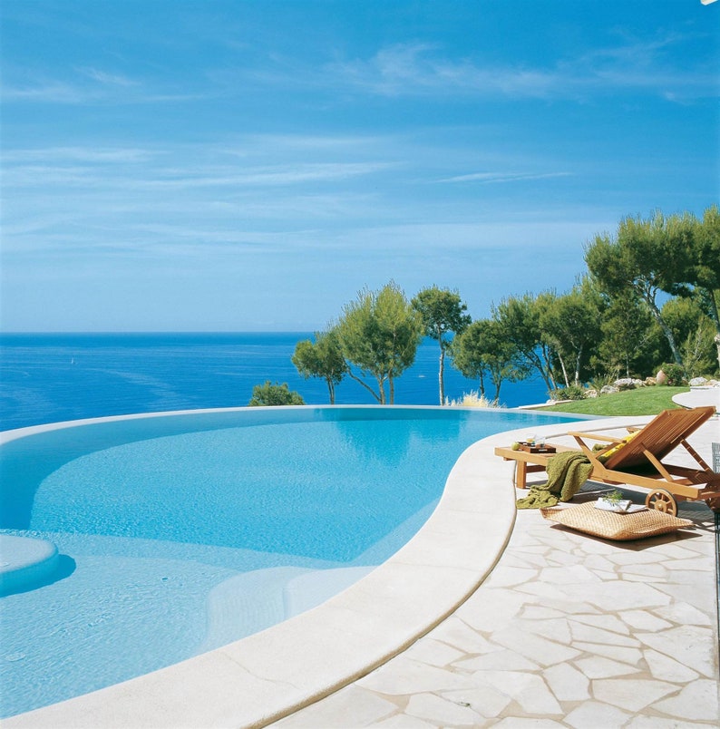 May include: A swimming pool with a curved edge, overlooking a blue ocean with a clear sky. A wooden lounge chair sits by the pool, with a towel and a pillow.