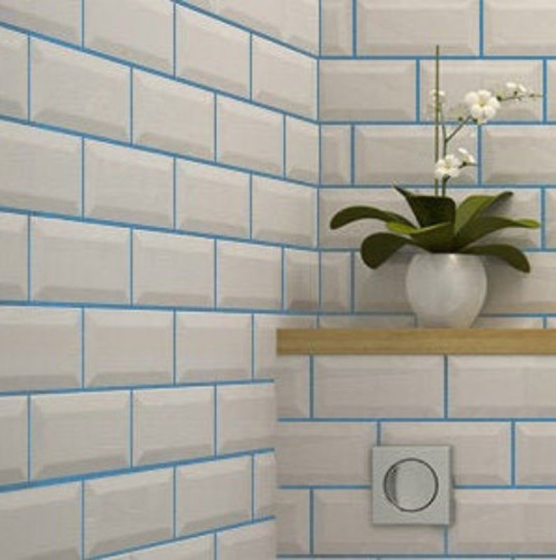 May include: White subway tile with blue grout lines. The tiles are arranged in a brick pattern. A small wooden shelf is mounted on the wall, with a white ceramic planter holding a green plant with white flowers.