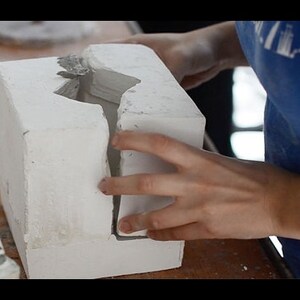 May include: A white plaster mold with a rough texture. The mold is open on one side, revealing a hollow space inside. The mold is being held by a person's hand.