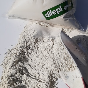 May include: A white bag of difepi grout powder is partially open, spilling out onto a white surface. A red and white trowel is lying on the powder.