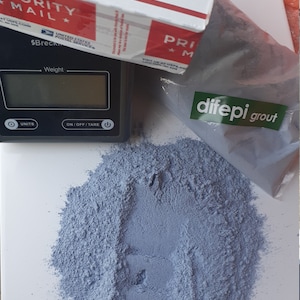 以下が含まれることがあります： 白い表面に「difepi grout」というラベルの付いた青い粉末のクローズアップ。粉末は緑色のラベルが付いた透明なビニール袋に入っています。背景にはデジタルスケールがあります。