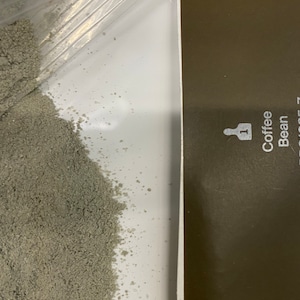 May include: A close-up of a white surface with a pile of grey coffee grounds. A brown surface with the text "Coffee Bean" and the number "1" is visible on the right side of the image.