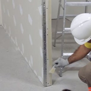 May include: A construction worker wearing a white hard hat and yellow shirt is using a tool to finish a wall. The wall is made of drywall and has a white finish. The worker is standing on a ladder and is using a tool to smooth out the drywall.