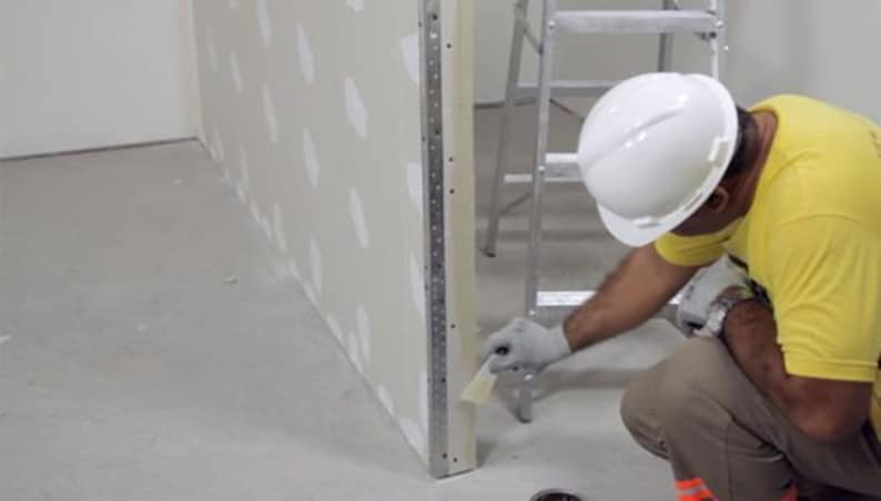 May include: A construction worker wearing a white hard hat and yellow shirt is using a tool to finish a wall. The wall is made of drywall and has a white finish. The worker is standing on a ladder and is using a tool to smooth out the drywall.