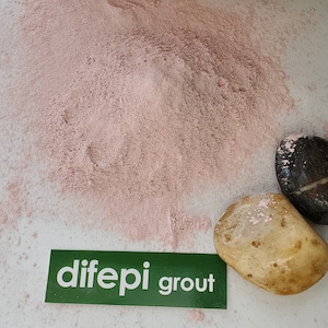 May include: A close-up of a pile of pink grout powder with two smooth stones on a white surface. The stones are covered in pink powder. A green sticker with the text "dife pi grout" is visible in the lower left corner of the image.