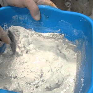 Puede incluir: Una mano enguantada mezcla una sustancia blanca y pulverulenta en una cubeta de plástico azul. La sustancia parece ser un tipo de yeso o cemento.