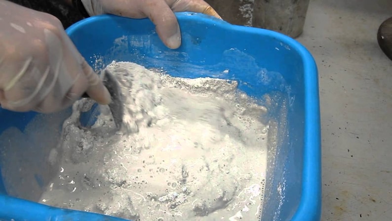May include: A gloved hand mixes a white, powdery substance in a blue plastic tub. The substance appears to be a type of plaster or cement.