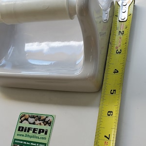 May include: A white ceramic toilet paper holder with a curved handle. The holder is approximately 8 inches long, as measured by a yellow tape measure. A sticker with the text "DiFEPI" is in the lower left corner.