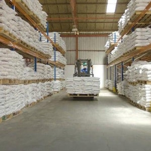 May include: A large warehouse with rows of white sacks stacked on shelves. A forklift is moving through the aisle, carrying more sacks.