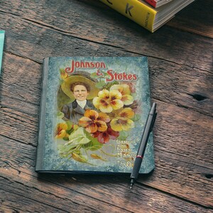 Puede incluir: Un cuaderno de estilo vintage con una cubierta azul que presenta a una mujer con un sombrero y un dise&ntilde;o floral. La cubierta dice "Johnson & Stokes" y "Garden, Farm and Flower Seeds 50c".
